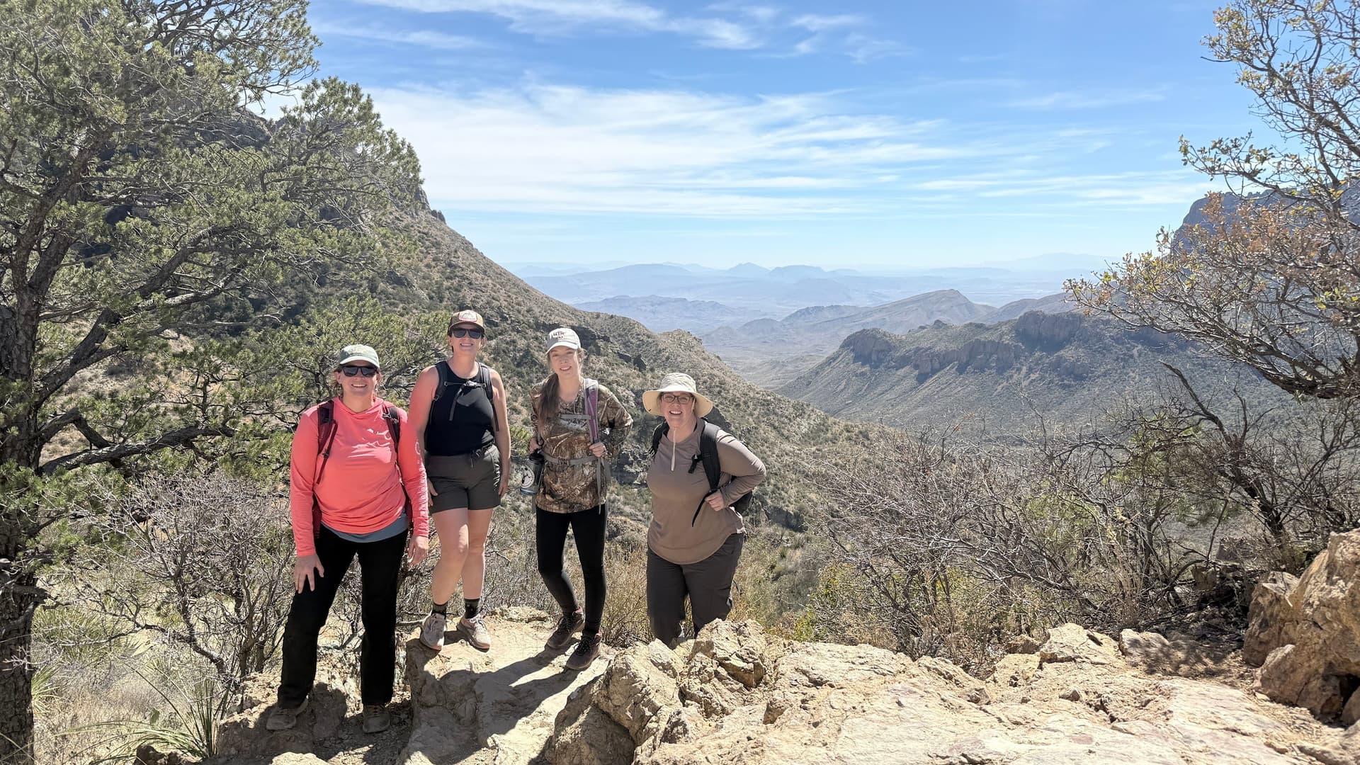  Four women hikers with backpacks on a rocky mountain trail overlooking the desert peaks of Big Bend, Texas