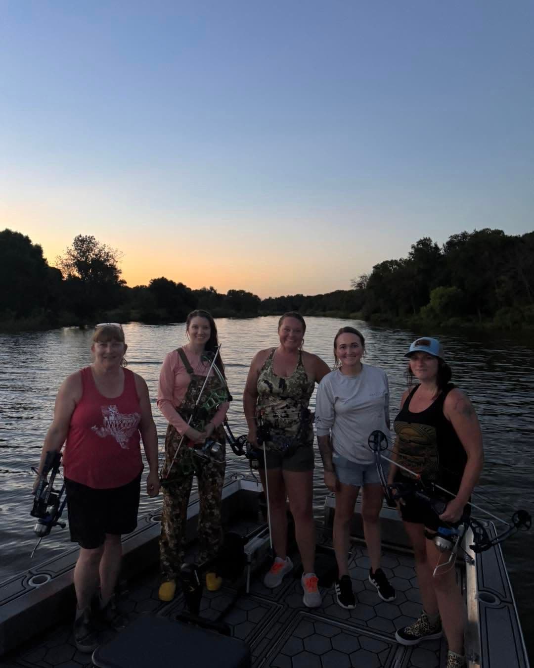  Five women holding bows on a flat-bottom boat during an evening bowfishing trip on a Texas river at dusk