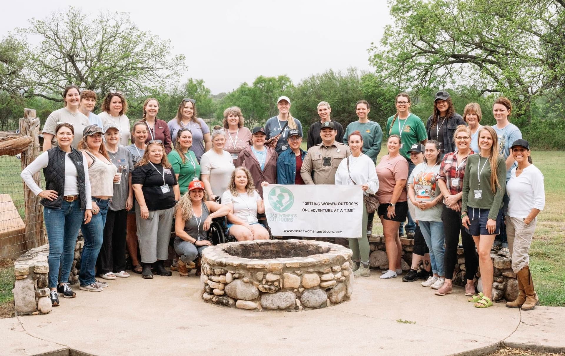 Large group of women around a stone fire pit holding a Texas Women's Outdoors banner at the 2026 annual retreat