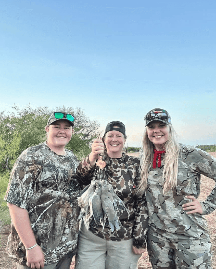 Three women in camouflage hunting gear smiling outdoors while holding a string of dove from a Texas hunt