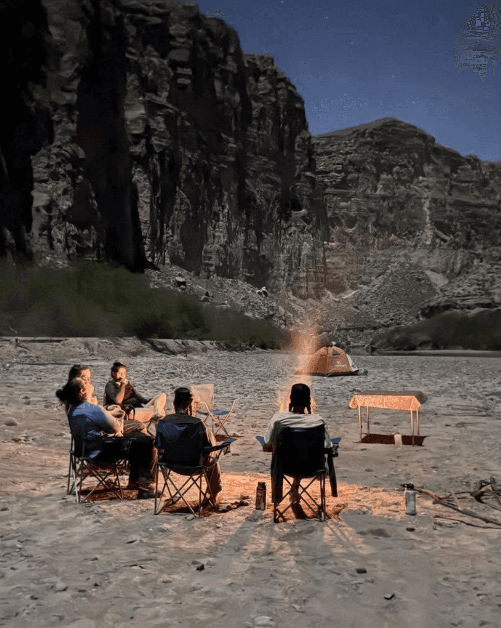 Group gathered around a campfire at night at a riverside canyon campsite in Big Bend, Texas, under a starry sky