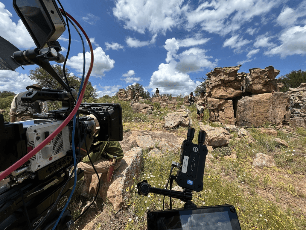 Film crew shooting a Western movie scene on a Texas ranch, with a cinema camera in the foreground and actors in period costume near a granite rock outcropping