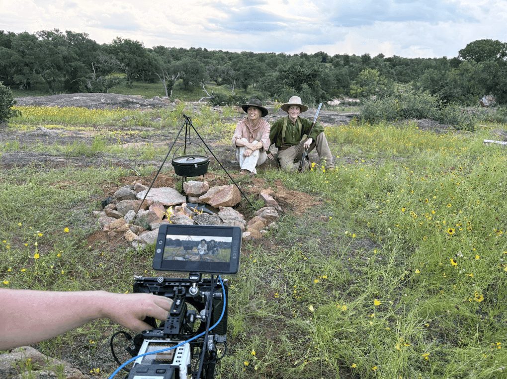 Two actors in Western costume sit by a campfire with a Dutch oven on a wildflower-covered Texas ranch, filmed on a handheld camera rig